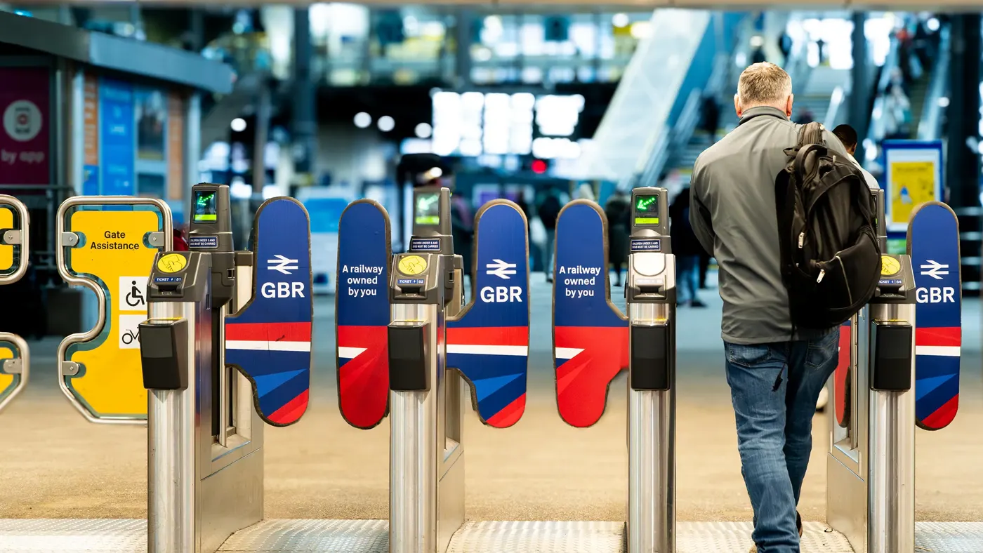 A rail passenger going through the ticket barrier at Leeds Train Station with the UK Government's re-nationalised rail company GBR signs and symbols displayed