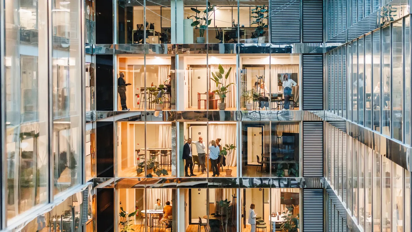 Men and women of various backgrounds collaborate and work independently in a spacious, well-lit office setting, each attired suitably for a professional setting, using modern tools and technology.