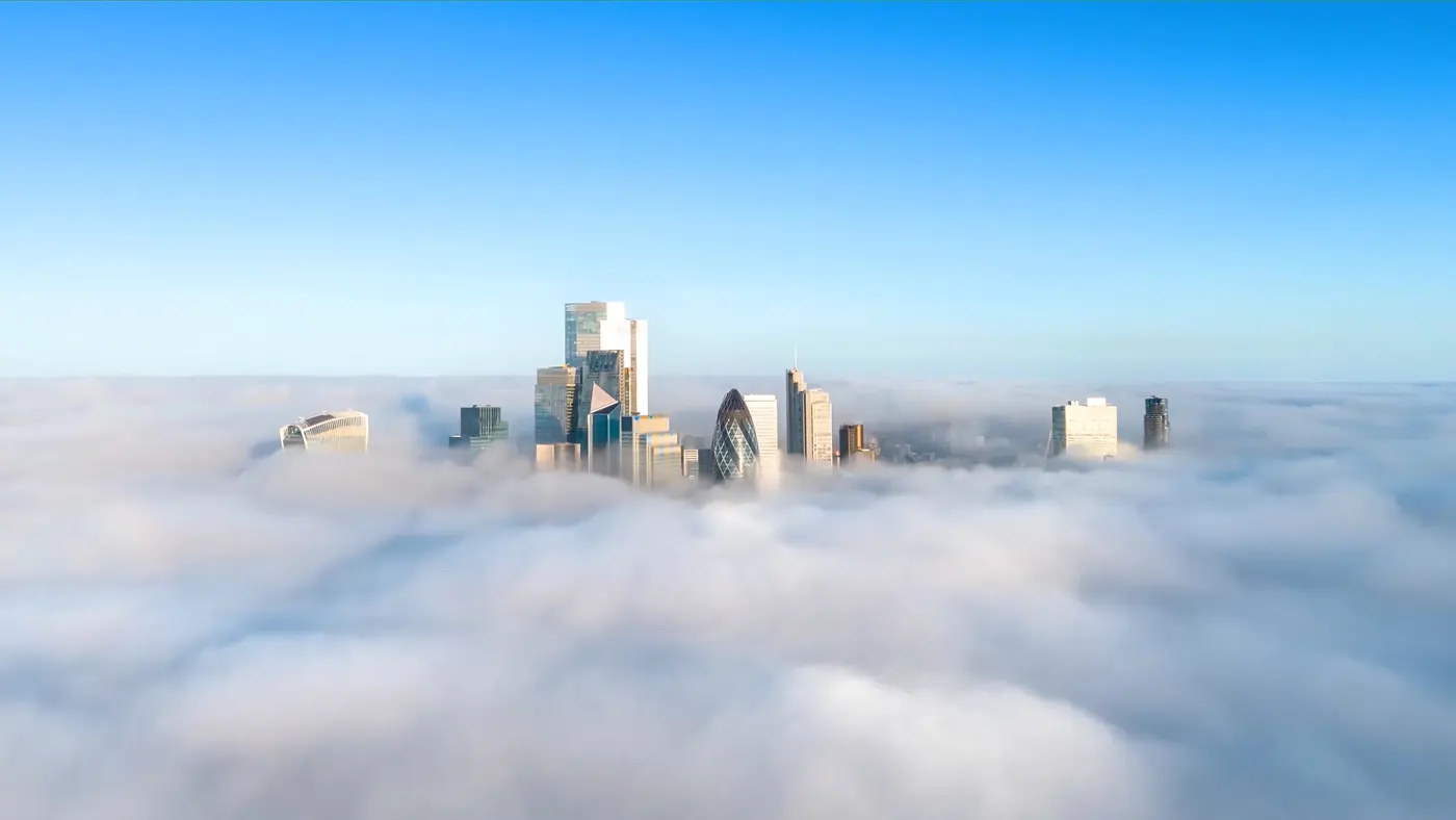 City skyline with modern skyscrappers rising above a layer of fog under a clear blue sky.