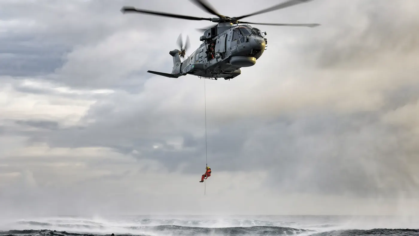 A crew member from a hovering military rescue helicopter is lowered by powered winch from a side door.