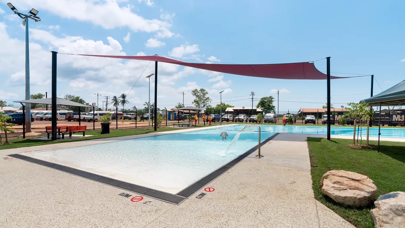 An outdoor swimming pool with shallow water, surrounded by a concrete deck. The pool has a sloped entry and a handrail on the right side. There are signs on the pool deck indicating "No Diving" and depth markings in both feet and meters. Above the pool, a large red sunshade is stretched between several poles to provide shade. The surrounding area includes some green grass, benches, and small trees. In the background, there are buildings, palm trees, and parked cars under a partly cloudy sky.