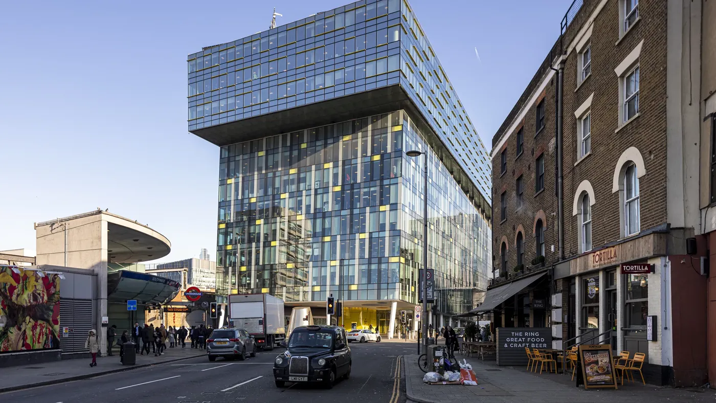 Modern urban street scene in London, featuring a striking contemporary glass building with a cantilevered upper section. This building is set against a backdrop of older brick structures, including a corner restaurant named "Tortilla" with outdoor seating. A classic black London taxi and other vehicles are visible on the street, along with pedestrians walking by. The area appears busy with a mix of architectural styles, reflecting both modern and historical elements of the city. 